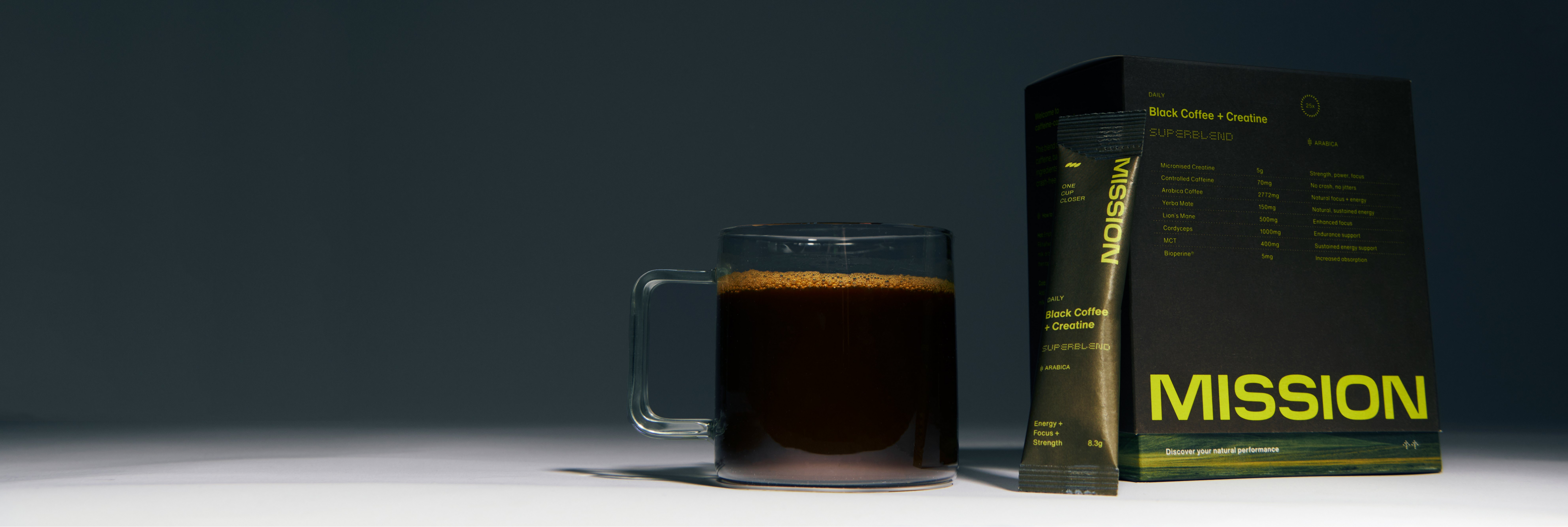 A glass mug filled with dark coffee sits beside a black Mission coffee sachet package with bright yellow-green text, photographed on a dark studio background.